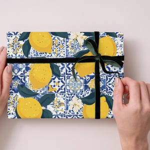 May include: A rectangular gift box with a lemon and tile pattern. The design features yellow lemons, green leaves, and blue and white tile motifs. A black ribbon and bow secure the package.
