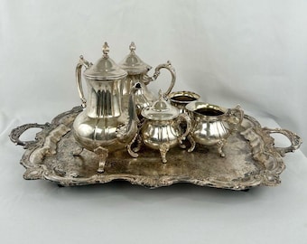 May include: A silver tea set on a decorative tray. The set includes a teapot, a creamer, a sugar bowl, and two cups. The tray has ornate handles and a detailed border. The tea set is reflective and appears to be made of silver.
