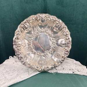 May include: Ornate silver-colored plate with a detailed, swirling floral design. The plate has a scalloped edge and a reflective center. The plate is likely vintage or antique, and is displayed on a green surface with a white lace doily.