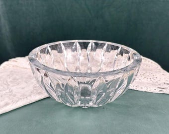 May include: A clear, cut-glass bowl with a scalloped rim and a repeating leaf-like pattern around the exterior. The bowl is round and sits on a green surface, with a white lace doily partially visible.