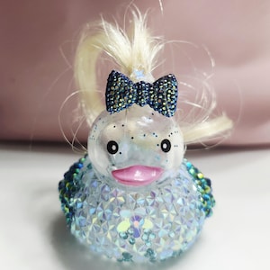 May include: A rubber duck figurine featuring a clear, iridescent body covered in multifaceted blue and silver crystals. The duck has a bright pink beak, black eyes, and a tuft of blonde hair topped with a dark blue, crystal-encrusted bow.
