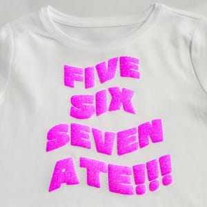 Könnte beinhalten: Weißes T-Shirt mit dem Schriftzug "FIVE SIX SEVEN ATE!!!" in leuchtend pinkfarbenen, stilisierten Buchstaben. Das Shirt hat einen Rundhalsausschnitt und kurze Ärmel. Der Text ist gestapelt angeordnet.