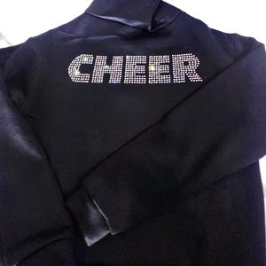 May include: A black long-sleeve sweatshirt featuring the word CHEER spelled out in clear, reflective rhinestones across the chest. The garment has a high neckline and ribbed cuffs, with the sparkling crystal lettering creating a bold contrast.