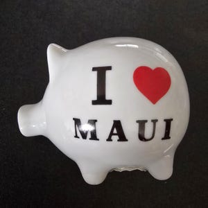 May include: White ceramic piggy bank shaped like a pig, with the text "I LOVE MAUI" in black letters and a red heart. The piggy bank has a slot on top for inserting coins.