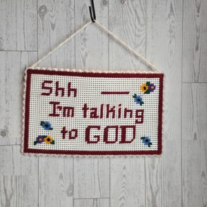 May include: A rectangular plastic canvas wall hanging featuring the maroon stitched text Shh — I'm talking to GOD. The piece is bordered in maroon with small blue, yellow, and purple floral accents and includes a white string for hanging.