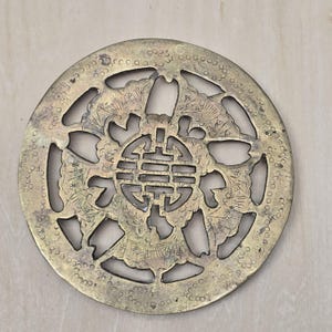 May include: A circular brass trivet featuring an intricate openwork design. The center displays a stylized character surrounded by radiating fan-shaped motifs and cloud-like patterns. The outer border is decorated with a repeating small circle pattern.
