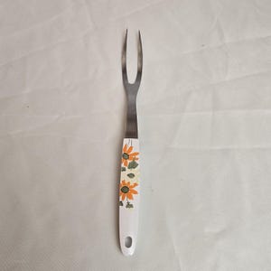 May include: A vintage carving fork with a stainless steel head and a white handle. The handle is decorated with orange, yellow, and green floral designs. The fork has two tines and a hole at the end of the handle for hanging.