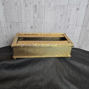 May include: A rectangular gold-colored box with a woven texture and a clear top. The box has a decorative border and small feet. The background is a light wood pattern and a black fabric.