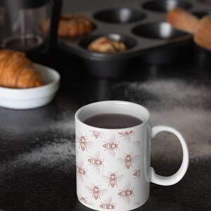 Może przedstawiać: Biały ceramiczny kubek z powtarzającym się wzorem małych, brązowych, szczeg&oacute;łowych ilustracji pszcz&oacute;ł. Kubek posiada standardowe ucho i jest wypełniony ciemnym płynem. Wz&oacute;r pokrywa całą zewnętrzną powierzchnię naczynia w r&oacute;wnomiernym układzie.