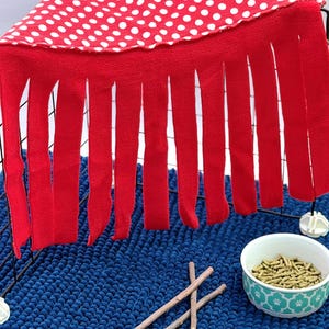 Puede incluir: Un refugio de tela roja con lunares blancos y flecos largos que cuelgan de un marco de alambre negro. Debajo del marco hay un pequeño cuenco redondo lleno de gránulos marrones y dos palos de madera finos sobre una alfombra azul oscuro texturizada.