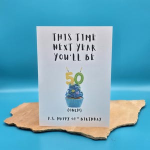 May include: A white card with the text "This time next year you'll be 50 (ouch)" and a blue cupcake with a 50 candle on top. The card also says "P.S. Happy 49th Birthday".