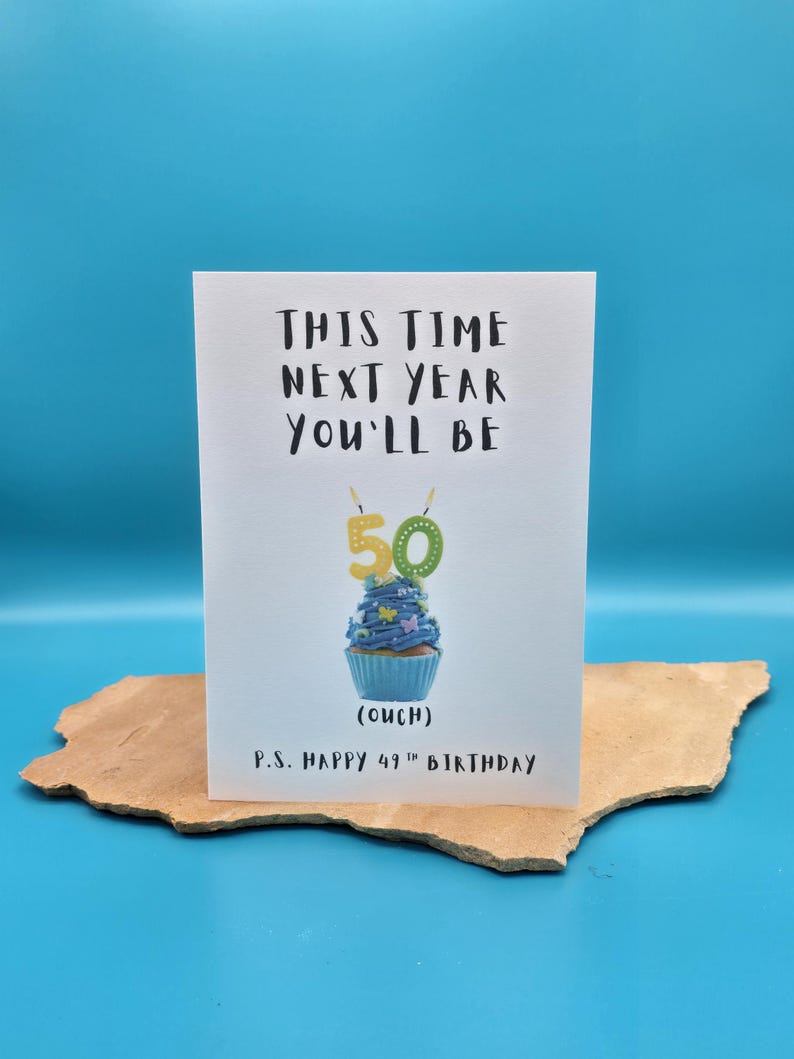 May include: A white card with the text "This time next year you'll be 50 (ouch)" and a blue cupcake with a 50 candle on top. The card also says "P.S. Happy 49th Birthday".