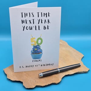 May include: A white card with black text that reads "This time next year you'll be 50 (ouch) P.S. Happy 49th Birthday". A blue cupcake with yellow candles that read "50" is pictured on the card.