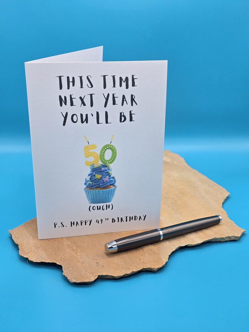 May include: A white card with black text that reads "This time next year you'll be 50 (ouch) P.S. Happy 49th Birthday". A blue cupcake with yellow candles that read "50" is pictured on the card.