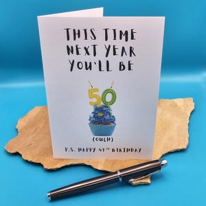 May include: A white card with black text that reads "This time next year you'll be 50 (ouch) P.S. Happy 49th Birthday". The card features a blue cupcake with a green and yellow number 50 candle.