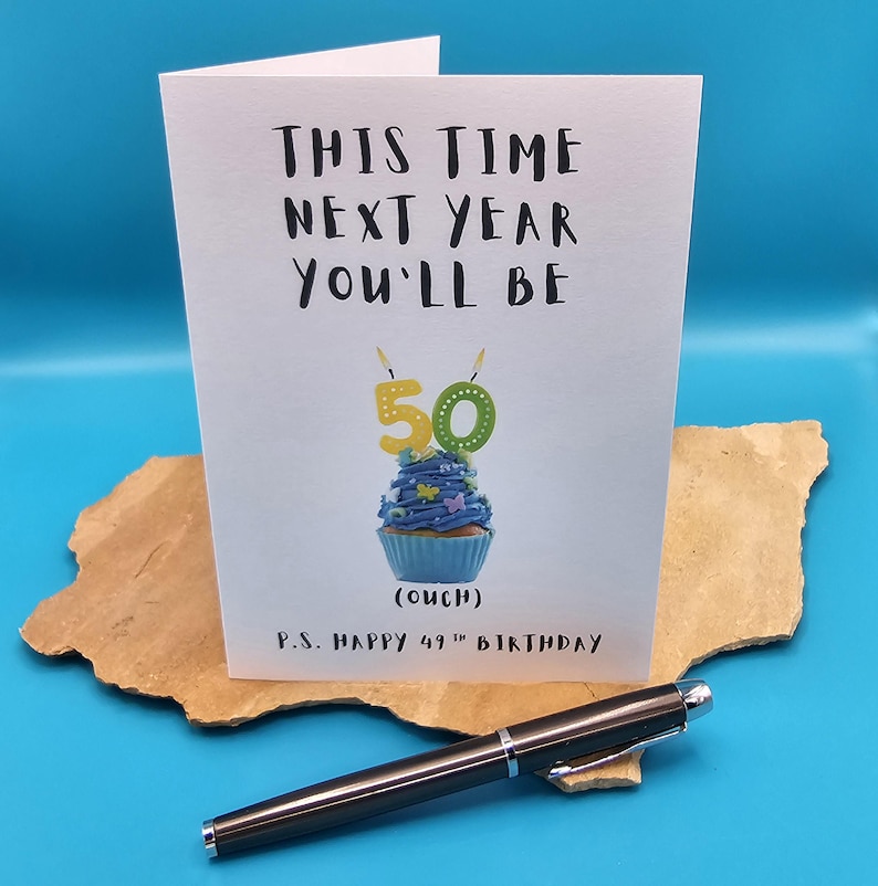 May include: A white card with black text that reads "This time next year you'll be 50 (ouch) P.S. Happy 49th Birthday". The card features a blue cupcake with a green and yellow number 50 candle.