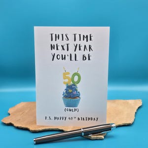 May include: A white card with black text that reads "This time next year you'll be 50 (ouch)".  A blue cupcake with yellow candles that read "50" is pictured on the card.  The text "P.S. Happy 49th Birthday" is at the bottom of the card.