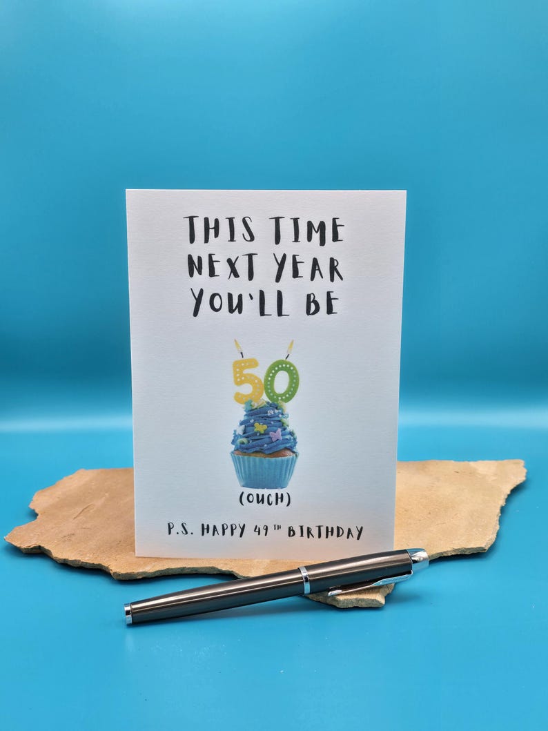 May include: A white card with black text that reads "This time next year you'll be 50 (ouch)".  A blue cupcake with yellow candles that read "50" is pictured on the card.  The text "P.S. Happy 49th Birthday" is at the bottom of the card.
