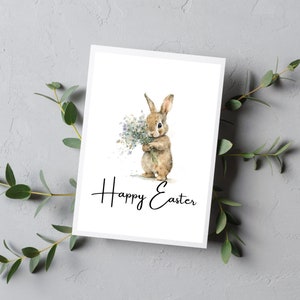 May include: A white card with a watercolor illustration of a brown bunny holding a bouquet of white flowers. The text "Happy Easter" is written in black cursive below the bunny.