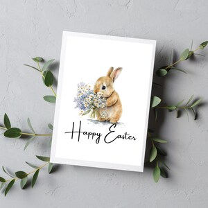 May include: A white card with a watercolor illustration of a brown bunny holding a bouquet of purple and white flowers. The text "Happy Easter" is written in black cursive below the bunny.
