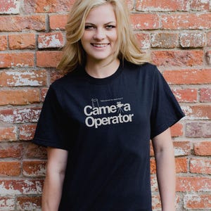Pode incluir: Uma camiseta preta com um gráfico branco que diz "Camera Operator" e inclui uma câmera e um tripé estilizados. O texto também inclui a frase "Quem dirigiu meu selfie de volta?"