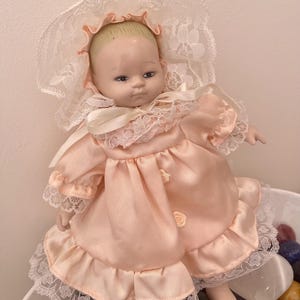 May include: A porcelain doll with blonde hair wearing a peach-coloured satin dress with lace trim and a matching bonnet. The dress features small fabric rosettes down the front, and the bonnet is adorned with white floral lace detailing.