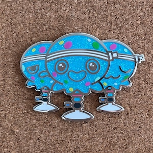 May include: A silver-outlined enamel pin featuring three cartoon jellyfish with smiling faces. The jellyfish are light blue with glitter and colorful polka dots. Each jellyfish has a white band around its head.