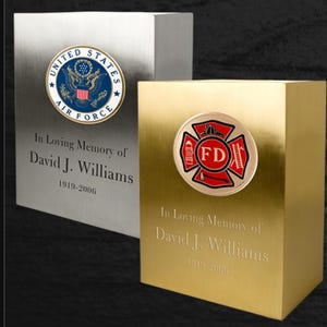 May include: Two silver and gold metal urns with engraved text. The silver urn has a United States Air Force emblem. The gold urn has a red and gold firefighter emblem. Both urns say "In Loving Memory of David J. Williams 1919-2006".