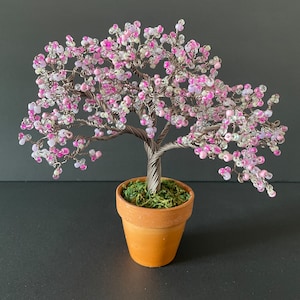 May include: A decorative wire tree sculpture with pink and white beaded flowers. The tree is in a small terracotta pot filled with green moss. The trunk and branches are made of twisted wire.