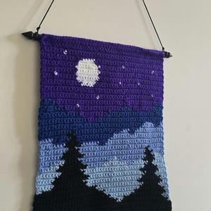 May include: A rectangular crochet wall hanging featuring a night landscape with a purple sky, a white moon, and small white stars. Below the sky are layers of blue mountains and black pine tree silhouettes. The piece hangs from a black wooden rod.