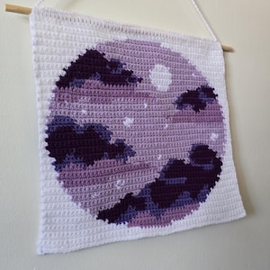 May include: A crocheted wall hanging with a night sky design. The artwork shows a purple and white night sky with a full moon and clouds. It is suspended from a wooden dowel with a white cord.