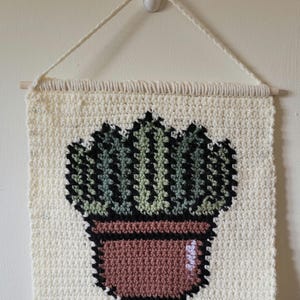 May include: A square crochet wall hanging featuring a potted cactus design. The background is off-white, with a brown pot and a green cactus outlined in black yarn. The piece is suspended from a wooden dowel with a matching off-white yarn hanger.