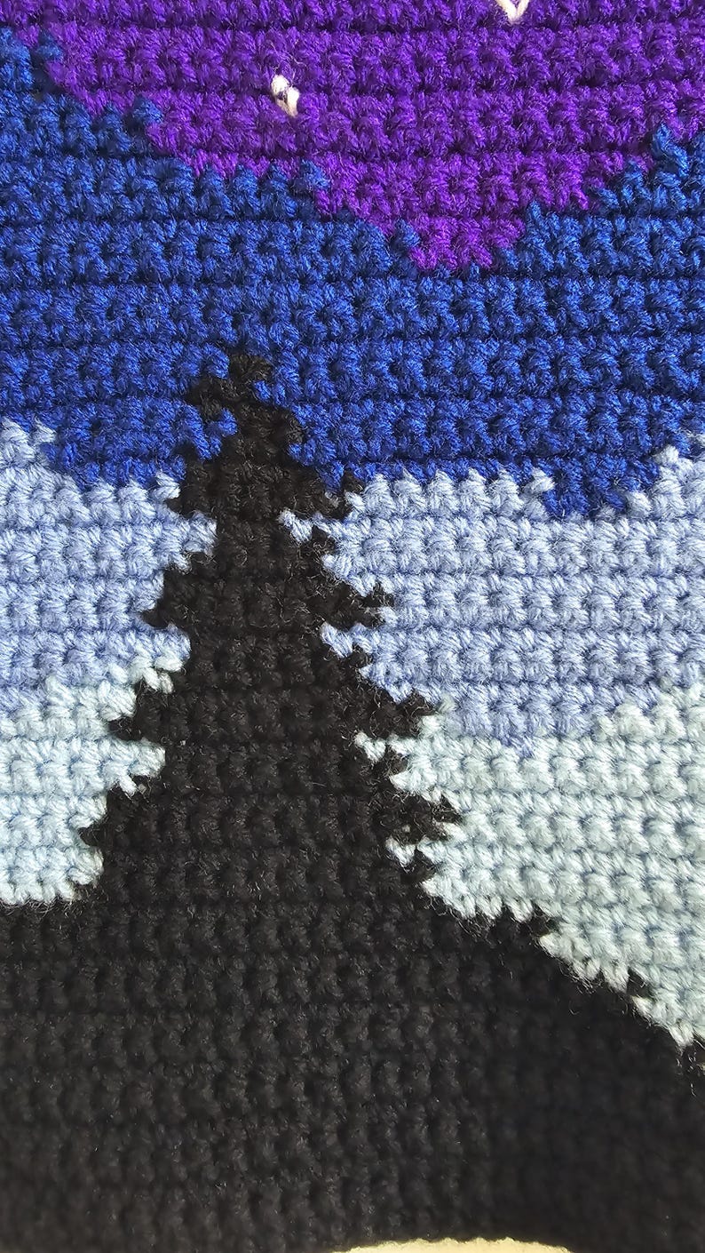 May include: A crochet tapestry featuring a landscape scene with a large black pine tree in the foreground. The background displays layers of light blue and medium blue yarn, topped with a dark purple sky containing small white stitched stars.