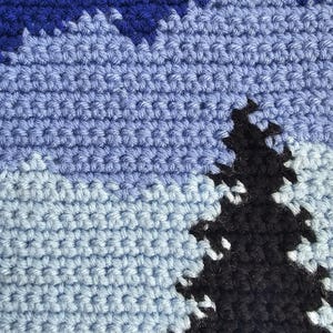May include: A crochet textile featuring a landscape design with a dark purple and blue sky, light blue mountains, and a prominent black evergreen tree silhouette in the foreground. The piece is constructed with visible yarn stitches in a dense pattern.