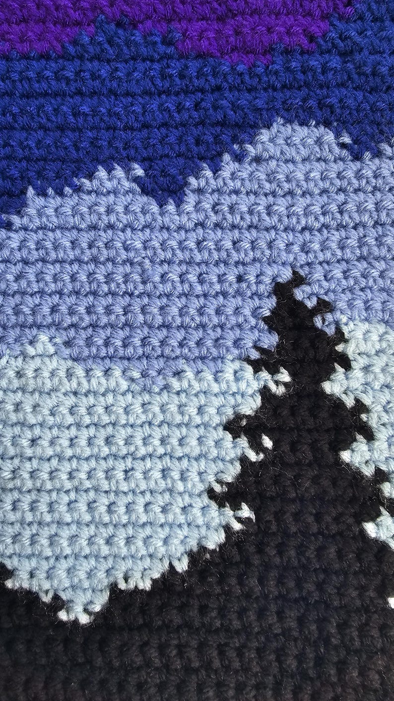 May include: A crochet textile featuring a landscape design with a dark purple and blue sky, light blue mountains, and a prominent black evergreen tree silhouette in the foreground. The piece is constructed with visible yarn stitches in a dense pattern.