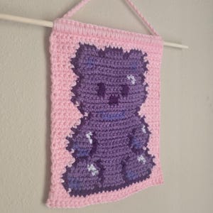 May include: A rectangular crochet wall hanging featuring a dark purple gummy bear design on a light pink background. The piece is suspended from a wooden dowel by a matching pink crocheted cord, showcasing a textured yarn stitch pattern throughout.