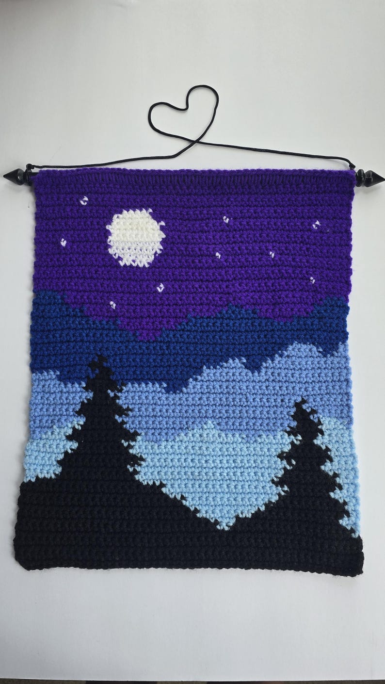 May include: A rectangular crochet wall hanging featuring a night landscape with a white moon and small white stars against a purple sky. Below the sky are layers of blue mountains and two large black pine trees in the foreground. A black cord hangs from a black rod.