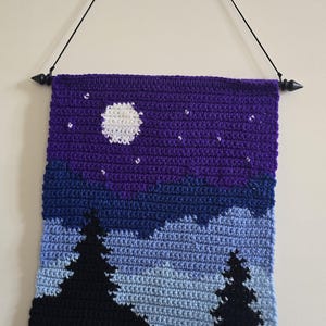 May include: A rectangular crochet wall hanging featuring a night landscape with a white moon and small white stars against a purple sky, layered over dark blue and light blue mountains, with two black pine tree silhouettes in the foreground.
