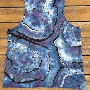 Pode incluir: Uma regata sem mangas com um padrão tie-dye em tons de azul, roxo e branco. O design apresenta formas orgânicas concêntricas com contornos pretos, criando um efeito marmorizado. A parte de cima é feita de tecido macio.