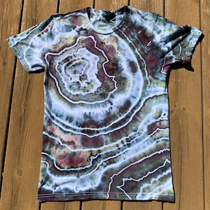 Puede incluir: Una camiseta tie-dye con un patrón circular en tonos de azul, verde, marrón y blanco. El patrón se asemeja a un anillo de árbol o a una ágata.