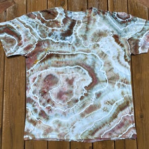 Op de afbeelding: Een T-shirt met korte mouwen met een tie-dye patroon in tinten bruin, wit en lichtblauw. Het ontwerp heeft concentrische cirkelvormige patronen, wat een unieke en artistieke uitstraling creëert. Het shirt is gemaakt van een zacht materiaal.