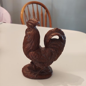 Puede incluir: Una figurita de gallo de madera con un acabado marrón. El gallo está de pie sobre una base con las plumas de la cola enroscadas.