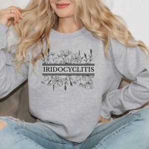 Puede incluir: Sudadera gris con un diseño floral y el texto "IRIDOCYCLITIS" impreso en la parte delantera.