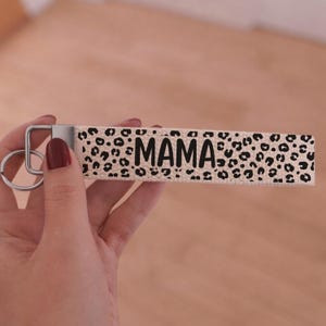 Può includere: Un portachiavi da polso beige con un motivo leopardato nero e la scritta "MAMA" in nero. Il portachiavi ha un anello e un fermaglio in metallo argentato.