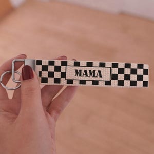 May include: A keychain wristlet with a black and white checkered pattern and the word "MAMA" in black block letters. The wristlet has a silver-colored metal key ring attached.