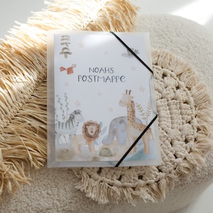 Könnte beinhalten: Ein weißer Ordner mit einem schwarzen Hefter. Die Vorderseite des Ordners zeigt eine Aquarell-Illustration von Tieren, darunter ein Zebra, ein Löwe, ein Elefant und eine Giraffe. Der Text "Noahs Postmappe" ist in Schwarz auf dem Ordner gedruckt.