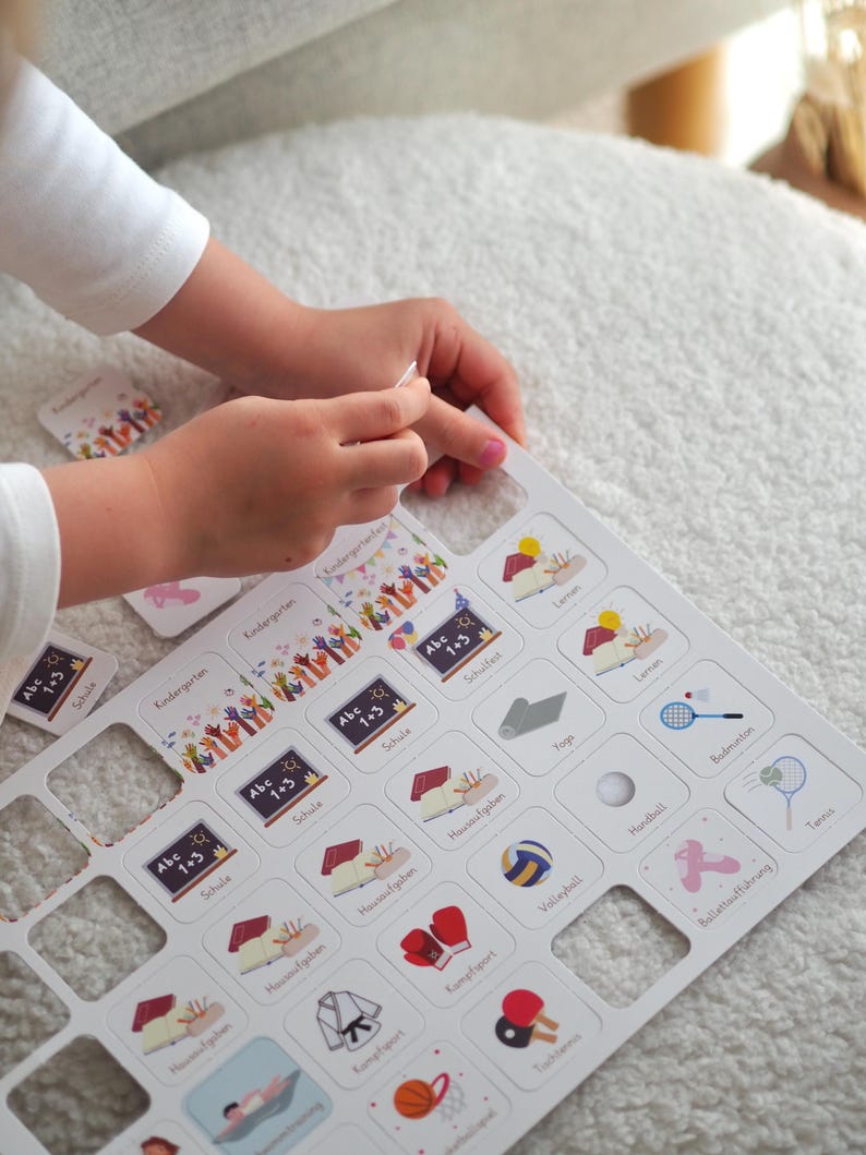 May include: A white board with a grid of squares, each containing an image and text. The images depict various activities, such as school, sports, and hobbies. The text includes words like "Kindergarten", "Schule", "Hausaufgaben", "Yoga", "Kampfsport", "Volleyball", "Handball", "Badminton", "Tennis", and "Ballett/Tanz/Figuren".