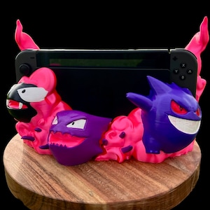 May include: A pink and purple 3D printed Nintendo Switch stand featuring the Pokemon Ghost-type characters Gengar and Haunter. The stand is on a wooden base.