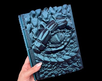 May include: A teal green leather-bound journal with a dragon eye and scales embossed on the cover.