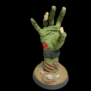 May include: A green zombie hand sculpture with red blood and a black wristband. The hand is reaching up and is on a brown base with rocks and bugs.